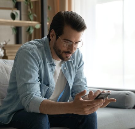 Serious millennial man in glasses sit on sofa holding smartphone check online calendar, sending message on messenger answers to friend, using business mobile app. Modern tech usage, leisure concept